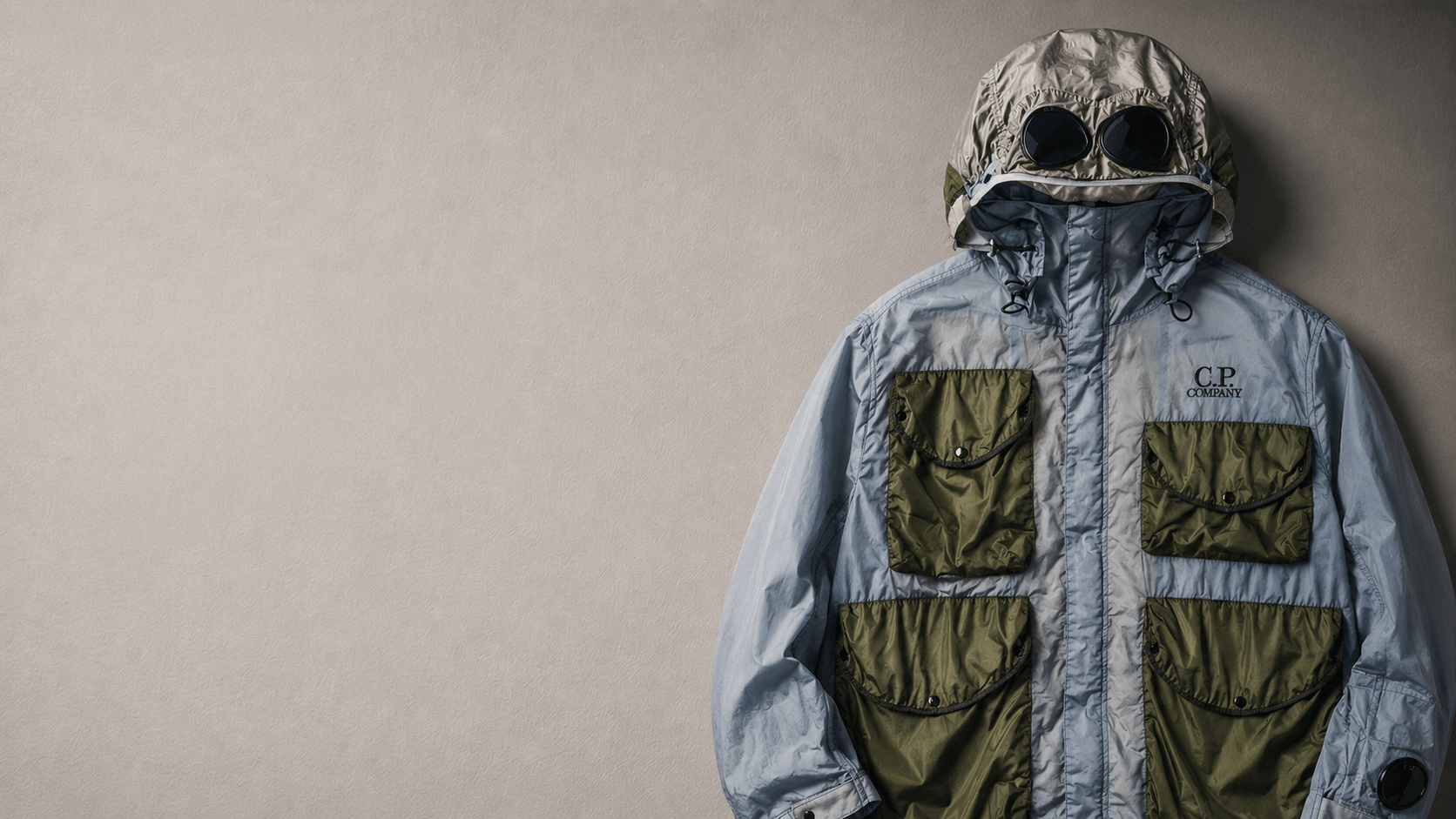 Two archive jackets hanging side by side on a plain warm wall: a Stone Island shell in yellow, green and black, and an olive C.P. Company goggle jacket